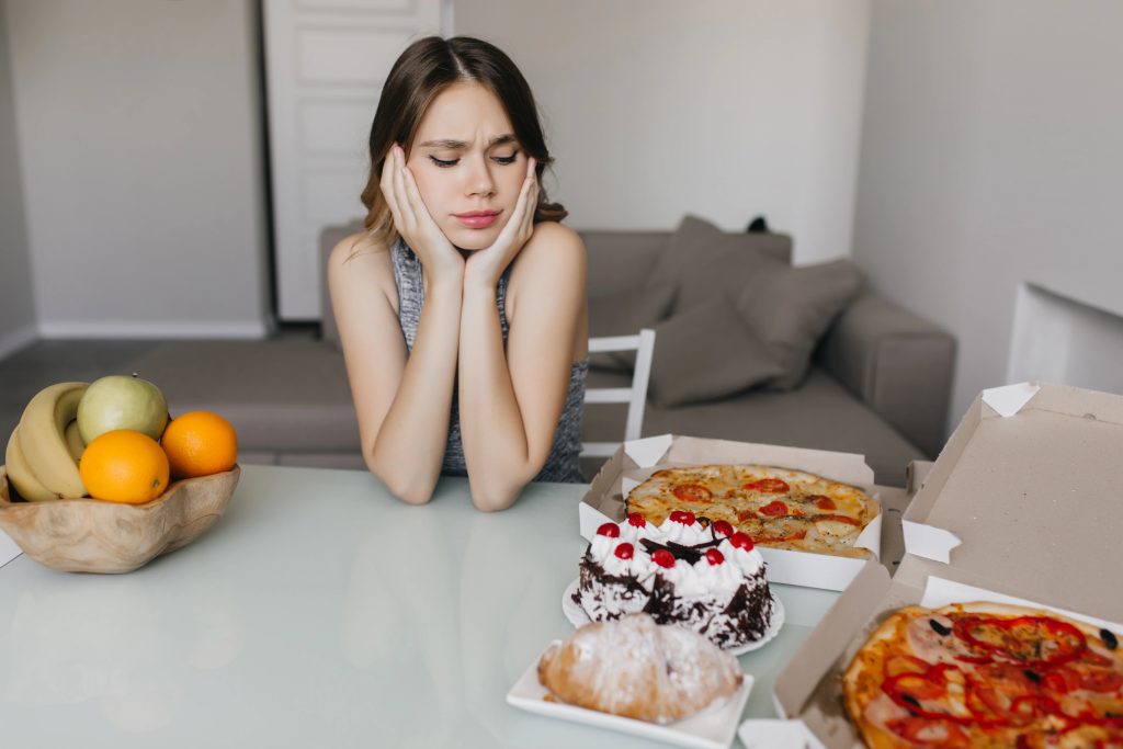 Young woman sitting at a table looking conflicted between healthy fruits and unhealthy foods like pizza and desserts.