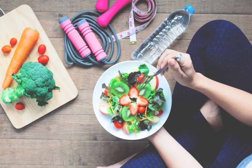 Top view of a person eating a fresh fruit and vegetable salad surrounded by healthy foods, water bottle, jump rope, and dumbbells, emphasizing a healthy lifestyle.